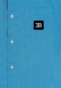 Camicia azzurro chiaro con una texture liscia, dotata di bottoni bianchi e una tasca nera con logo ricamato bianco "EB."
