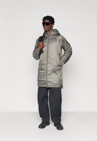 Hosszú, fémes szürke puffernadrág kabát kapucnival, cipzárral és oldalsó zsebekkel, sötét nadrággal és fekete cipővel párosítva. A modellen napszemüveg van.