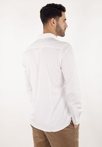 Hvit skjorte med lange ermer og spiss krage, glatt tekstur og mansjetter med knapper, brukt sammen med khakifargede kordfløyelsbukser, sett bakfra.