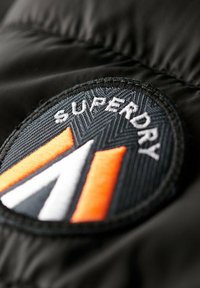 Zwarte stof met een ronde patch met de tekst "SUPERDRY", oranje en witte chevronaccenten en een gestructureerde achtergrond.