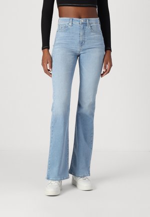 Kvinde iført lyseblå højttaljede flare jeans, hvide sneakers og en sort cropped langærmet top mod en ensfarvet baggrund.