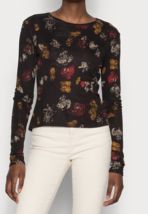 Musta pitkähihainen paita, jossa on abstrakteja punaisen, keltaisen ja valkoisen kukkakuoseja, päällä seisovalla henkilöllä, joka käyttää off-white-housuja.