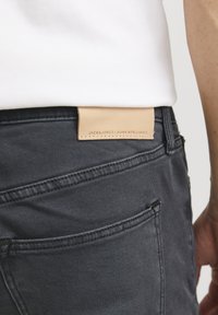 Tamnosivi traperici s kožnom etiketom na pojasu koja prikazuje "JACK & JONES / JEANS INTELLIGENCE." Ima standardne stražnje džepove.