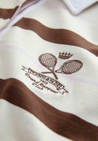Tissu rayé crème et marron avec des raquettes de tennis brodées, une couronne et le texte "INTEGRALITY The Champions Club."