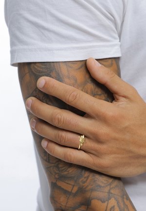 Main avec une bague en fil barbelé doré reposant sur un avant-bras tatoué, portant une chemise blanche à manches courtes.