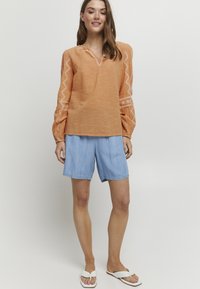 Orange linned blouse med broderede hvide mønstre, med V-hals og puffede ærmer, parret med lyseblå denim shorts. Hvide sandaler.