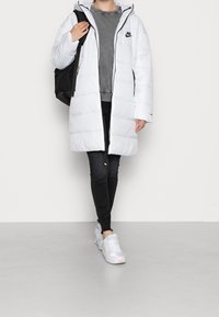 Personne portant un long manteau d'hiver Nike blanc, un jean noir, des baskets blanches, un sweat-shirt gris, portant un sac à dos noir.