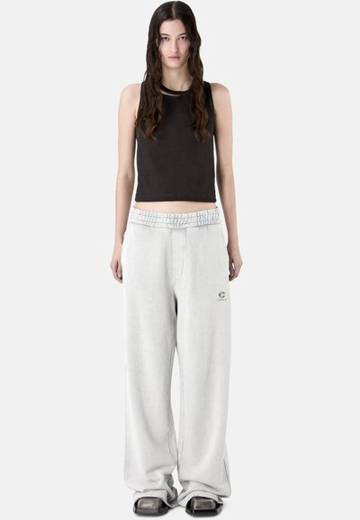 Femme aux longs cheveux foncés portant un crop top noir sans manches, un pantalon de survêtement blanc ample avec logo, et des chaussures noires à bout carré, debout droite.