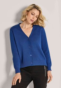 Vrouw met kort blond haar, gekleed in een gestructureerde blauwe knoopcardigan en zwarte broek met een zijrits, staande tegen een neutrale achtergrond.