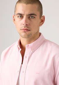 Világos rózsaszín gombos ing, gallérral, fehér gombokkal és egy kis Levi's logóval a mellkason. Puha anyagból készült, sima textúrával.