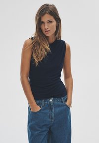 Navy sleeveless top with a draped design, paired with high-waisted blue denim jeans. Simple and smooth textures, casual fit.