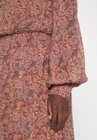 Main à la peau foncée reposant sur le côté, portant une robe à manches longues avec un motif floral couleur rouille, une taille élastique et des poignets de manches froncés.