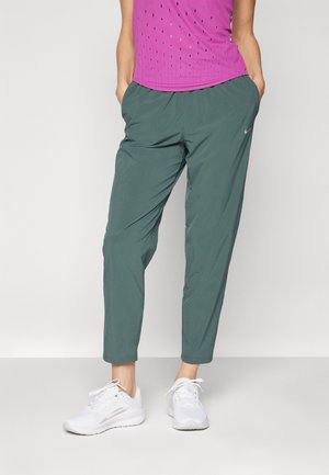 Osoba w ciemnozielonych sportowych spodniach, jasnoróżowej perforowanej bluzce i białych butach do biegania, stojąca z rękami w kieszeniach.