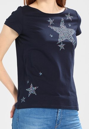 Femme portant un t-shirt bleu marine à manches courtes avec des motifs d’étoiles florales multicolores, associé à un jean en denim bleu.