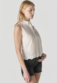 Blonde vrouw die een mouwloze witte blouse met borduursels en knopen draagt en zwarte denim shorts, staand in profiel tegen een grijze achtergrond.