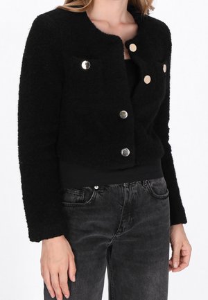 Vrouw die een korte zwarte textuurige jas met zilveren knopen draagt over een zwart shirt en vervaagde zwarte spijkerbroek, staand tegen een witte achtergrond.