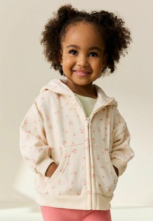 Roomkleurige zip-up hoodie met kleine roze bloemen, voorzien van een kangoeroezak en geribbelde manchetten, gedragen over een lichtgroene top.