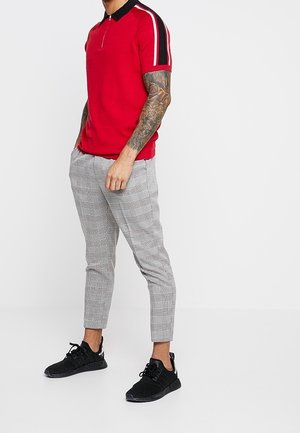 Polo rouge avec col noir et manches rayées, associé à un pantalon fuselé à carreaux gris et des chaussures de sport noires.