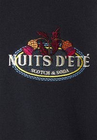 Tela negra con "Nuits d'Été" y "Scotch & Soda" bordados en blanco y motivos florales coloridos. Textura suave, diseño redondo.