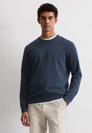 Marineblauer Sweatshirt aus weichem Baumwollstoff, mit Rundhalsausschnitt und gerippten Bündchen. Über einem hellen Hemd getragen, mit entspannter Passform.