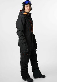 Zwarte sneeuwoverall met oranje accenten, ritszakken, geïsoleerde handschoenen en skibril. Het materiaal lijkt duurzaam en weersbestendig.