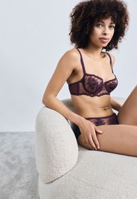 Lingerie set featuring sheer purple fabric with floral embroidery, intricate lace trim, and adjustable straps; model seated on a textured white chair.