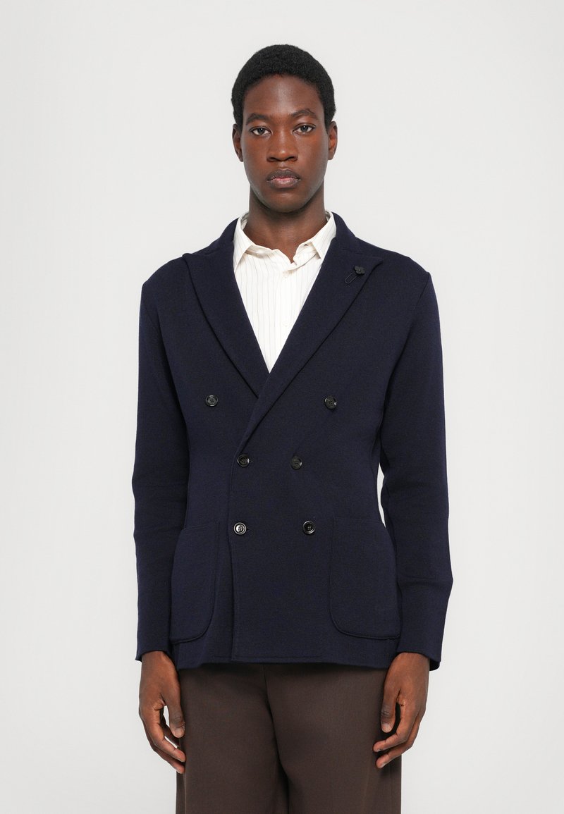 Navy blue double-breasted jacket in textured fabric, featuring black buttons, two front pockets, and a shawl collar layered over a white shirt.