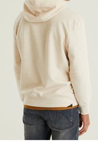 Sweat à capuche beige en tissu texturé, avec capuche et poche kangourou, superposé à une chemise marron, associé à un jean bleu foncé.