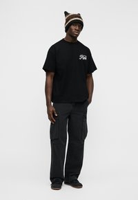 Άνδρας που φοράει μαύρο μπλουζάκι με μικρό λευκό κείμενο, μαύρο cargo παντελόνι, μαύρα παπούτσια και πολύχρωμο ριγωτό πλεκτό καπέλο με αυτιά.