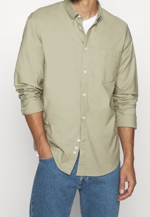 Mann trägt ein hellolivfarbenes Hemd mit Knöpfen über einem weißen T-Shirt mit hochgekrempelten Ärmeln, kombiniert mit blauen Jeans, Hände locker an den Seiten.