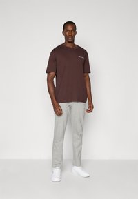 Brun kortærmet t-shirt med logo-detalje, parret med lysegrå sweatpants. Modellen står foran en ensfarvet baggrund iført hvide sneakers.