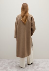 Cappotto lungo in lana marrone con una vestibilità comoda, spalle scese e fessure laterali, abbinato a pantaloni ampi beige. Texture morbida e design minimale.