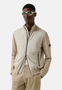 Beige bomber jacket with a smooth fabric front and knit sleeves. Features a high collar, zipped front, and two side pockets. Worn with light sunglasses.