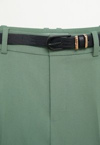 Ceinture noire texturée avec boucles dorées attachée autour de la taille d’un pantalon vert avec passants de ceinture et détail de coutures à l'avant.