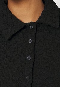 Black textured button-up shirt with a collar, featuring five rounded black buttons, and a crinkled fabric pattern.