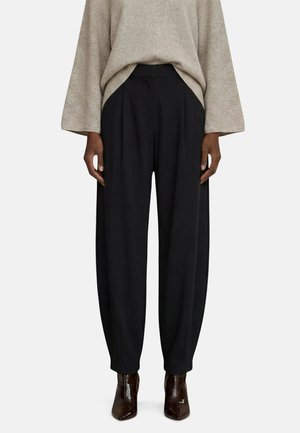 Personne portant un pull beige à manches longues, un pantalon plissé noir taille haute et des bottines marron foncé à bouts pointus.