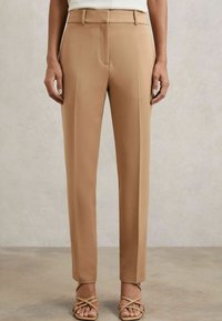Pantaloni beige su misura con un design a gamba dritta, dettaglio delle pieghe e tasche frontali, abbinati a tacchi a spillo leggeri. Tessuto dalla texture liscia.