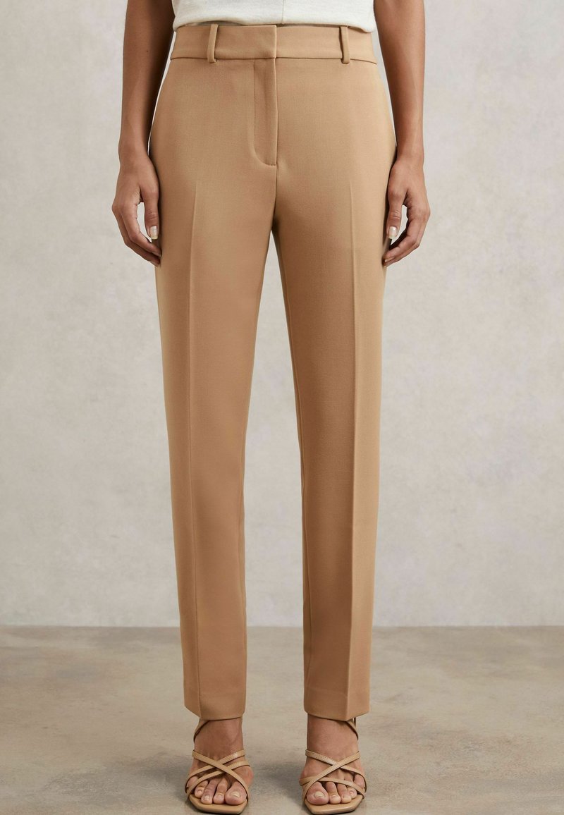 Pantaloni beige su misura con un design a gamba dritta, dettaglio delle pieghe e tasche frontali, abbinati a tacchi a spillo leggeri. Tessuto dalla texture liscia.