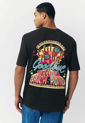 Sort t-shirt med farverig grafisk tekst og abstrakt design på ryggen, der fremhæver "Elton John Goodbye Yellow Brick Road."