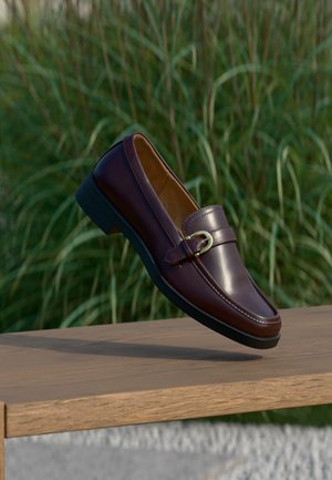 Mocassin en cuir marron foncé avec détail de boucle dorée suspendu au-dessus d’un banc en bois, fond d’herbe verte.