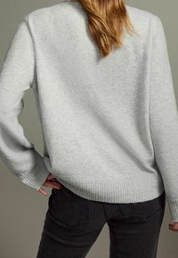 Pull en maille gris clair avec ourlet et poignets côtelés, présentant une coupe décontractée et une texture douce, porté avec un jean noir foncé.