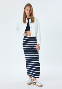 Witte cropped jacket, marineblauwe gestreepte maxi rok en sneakers met rubberen zolen. Jacket heeft drukknoopsluitingen; rok heeft een elastische tailleband.
