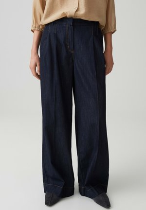 Beige blouse ingestopt in hooggesneden, wijde donkerdenim jeans, gedragen met puntige zwarte schoenen, staand tegen een lichte achtergrond.