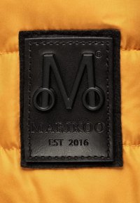 Black leather patch with raised "M" and circular accents, stitched to an orange fabric background; text reads "MARIKOO EST 2016."