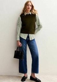 Green knitted vest over a white and green striped shirt, paired with wide-leg blue jeans and black loafers, holding a small black handbag.