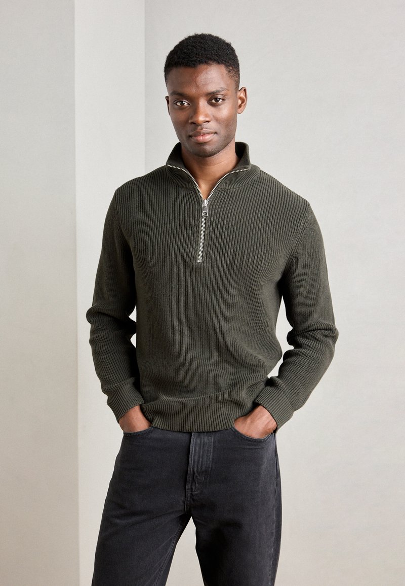 Suéter de punto con cuello de cremallera en verde oliva oscuro con una textura acanalada, combinado con jeans gris oscuro. Diseño simple, sin patrones ni acentos visibles.