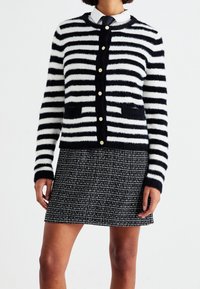 Svartvit randig cardigan med mjuk textur, utrustad med guldfärgade knappar och två framfickor. Bärs över en texturerad svart kjol.