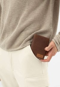 Bruine leren portemonnee met een geperforeerde textuur, gedeeltelijk zichtbaar in een witte zak, naast een lichtbeige gebreide trui.