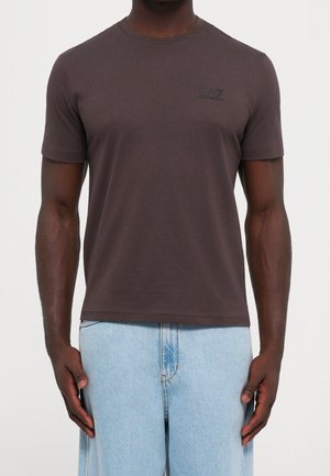 Homme portant un t-shirt brun foncé à manches courtes avec un petit logo sur la poitrine, associé à un jean bleu clair, debout devant un fond uni.