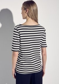 Camisa de manga corta a rayas blancas y negras con un escote amplio, confeccionada en tela suave y con un corte relajado, vista desde la parte trasera sobre un fondo neutro.